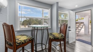 Un comedor con dos sillas de madera y una mesa de vidrio, ventanas amplias con persianas y vista a una escalera que lleva a un espacio exterior.