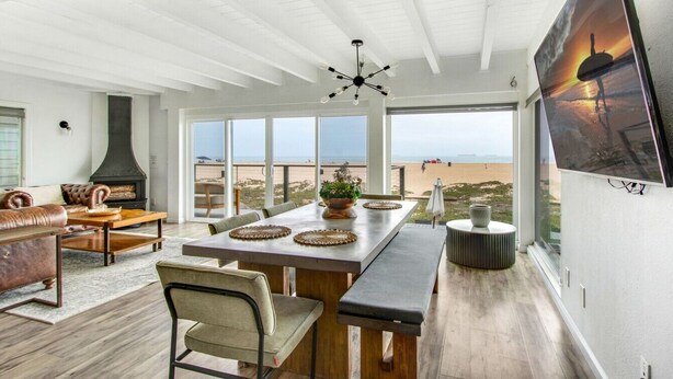 A spacious living area with a large window overlooking a beach, a fireplace, and a dining table with chairs. - Beachfront Sunset Beach, Ocean Views, Sleeps 10 (Huntington Beach)