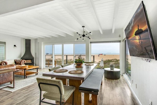 A spacious room with a large window overlooking a beach, a fireplace, and a dining area with a long table. - Beachfront Sunset Beach, Ocean Views, Sleeps 10 (Huntington Beach)
