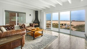 A living room with a leather sofa, a wooden coffee table, and a fireplace. There is a large window offering a view of the beach. - Beachfront Sunset Beach, Ocean Views, Sleeps 10 (Huntington Beach)