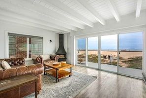 A living room with a leather sofa, a wooden coffee table, and a fireplace. There is a large window offering a view of the beach. - Beachfront Sunset Beach, Ocean Views, Sleeps 10 (Huntington Beach)