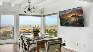 A dining area with a view of the beach, a large window, and a mounted TV. - Beachfront Sunset Beach, Ocean Views, Sleeps 10 (Huntington Beach)