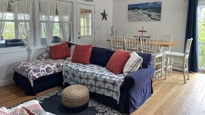 A cozy room with a wooden ceiling, a dining table with chairs, a sofa with patterned cushions, and a large window with sheer curtains. - Beach house Davis Park, Fire Island  (brookhaven)