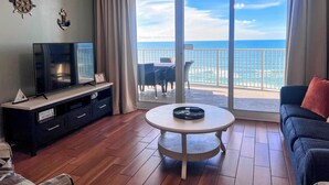 A living room with a TV, a small table, and a sofa. There is a large window with a view of the ocean. - BEACHFRONT Condo! FREE BEACH SERVICE! Sleeps 8! 2Bed 2Bath + Sleeper (Panama City Beach)