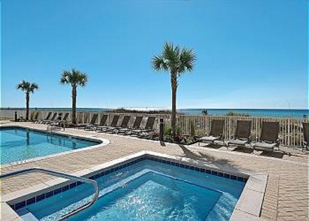 Ein Swimmingpool mit Liegestühlen und Palmen, mit Blick auf einen Strand.