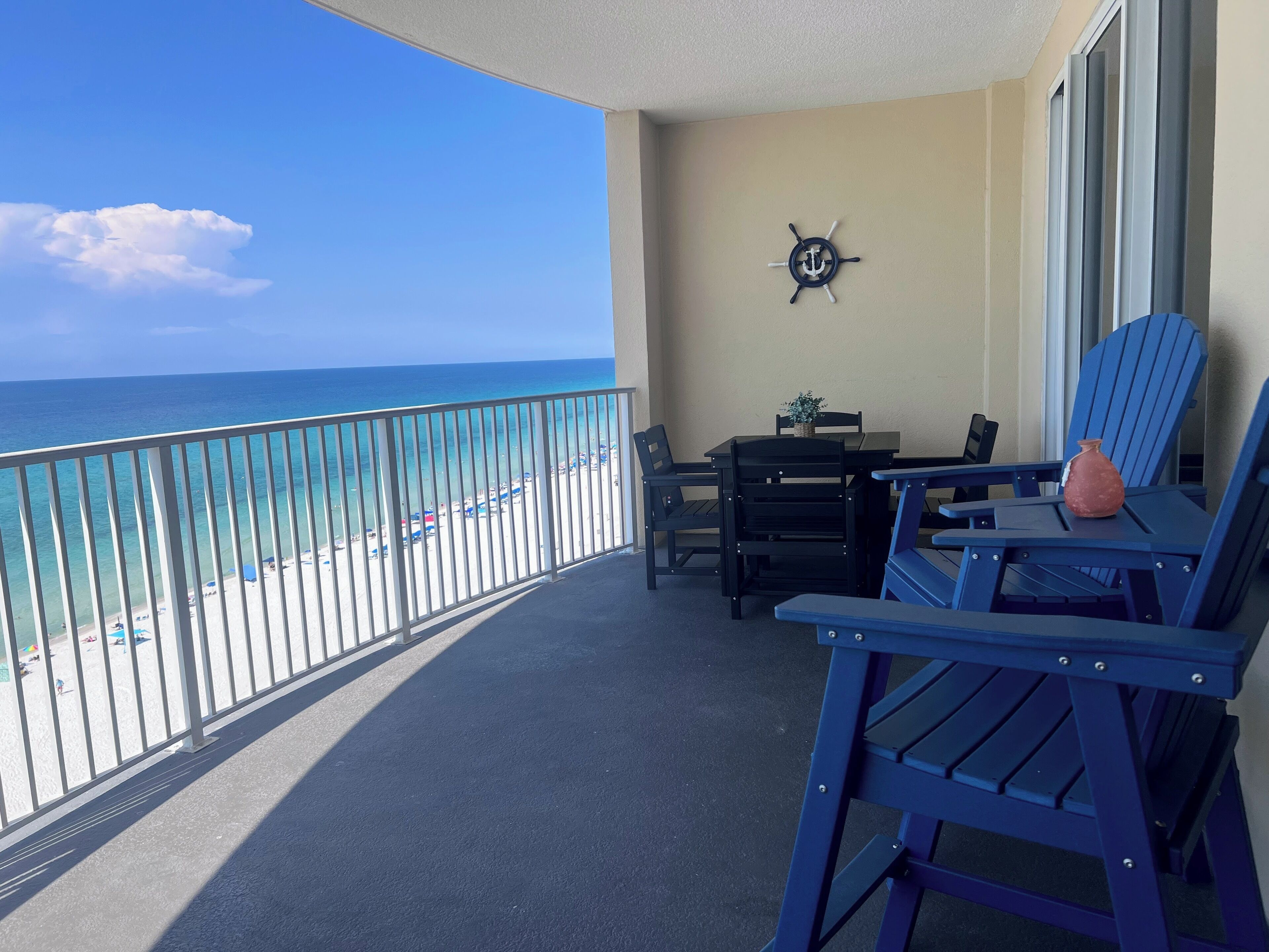 Een balkon met uitzicht op het strand en de oceaan. Er is een tafel met stoelen, een bloempot en een decoratieve ankerwandhanger.
