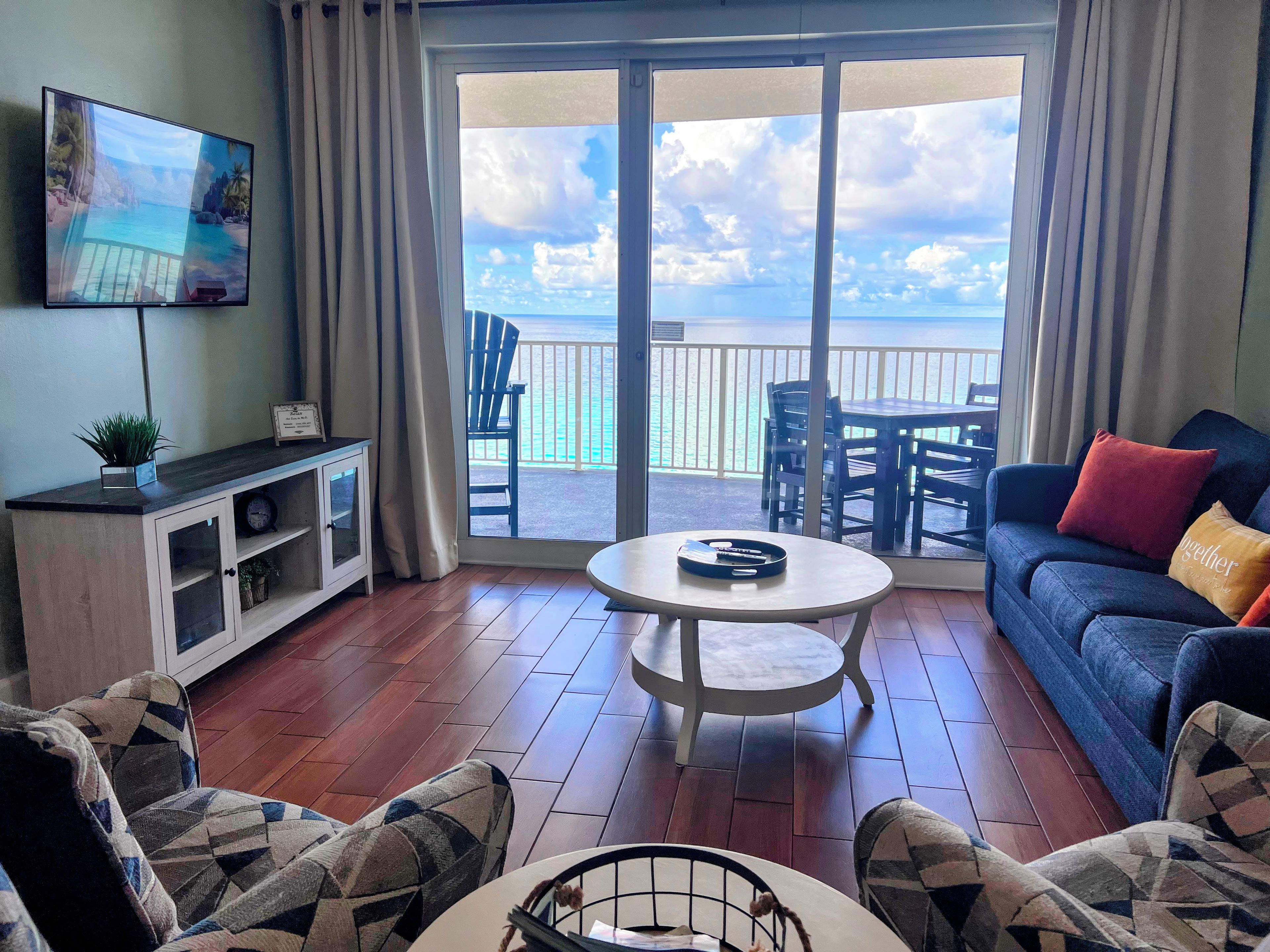 A living room with a TV, a sofa, a coffee table, patterned chairs, a TV stand, and a balcony with a view of the sea.