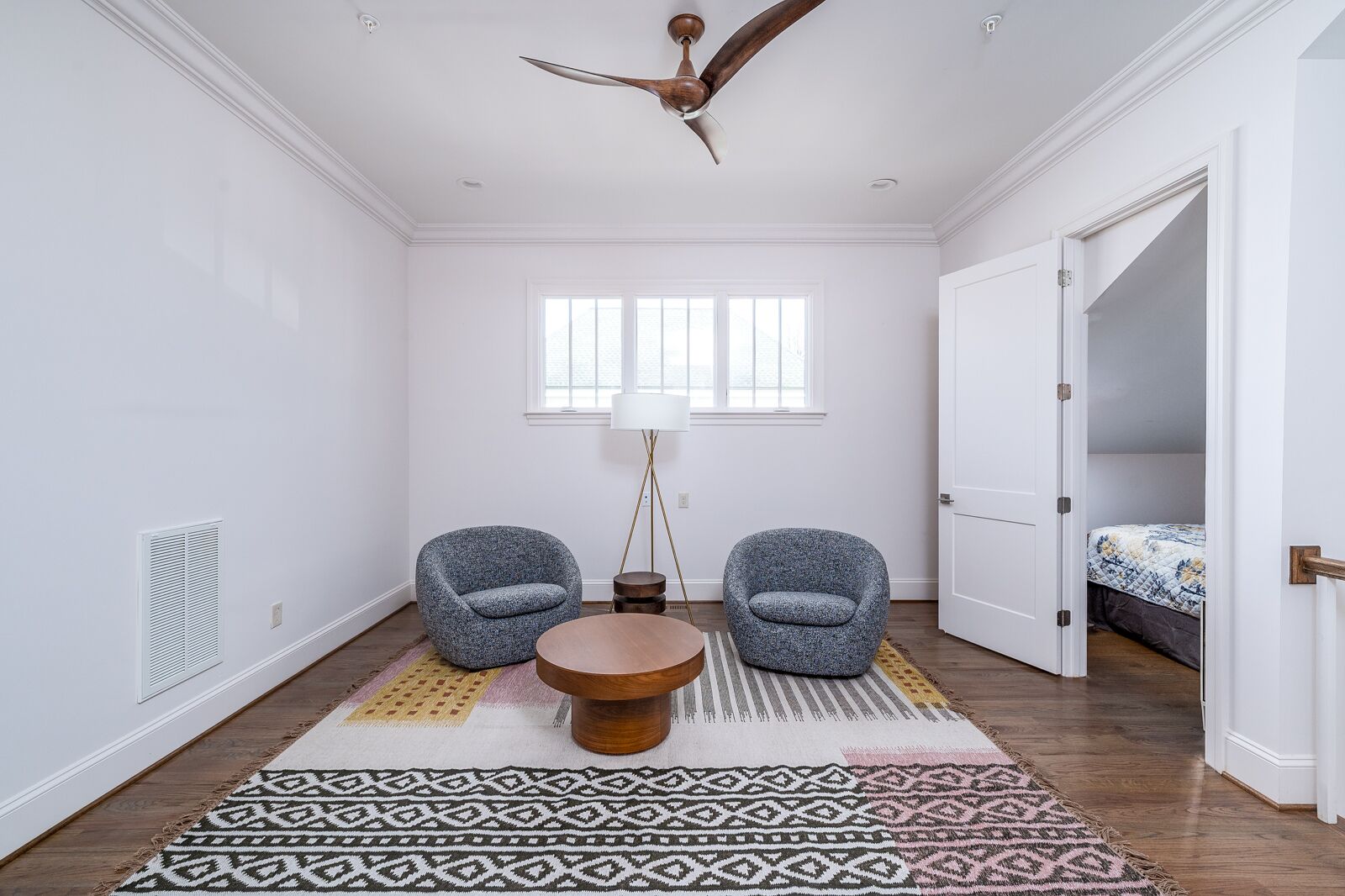 A spacious room with a high ceiling, two patterned armchairs, a round wooden table, a floor lamp, and a large window.