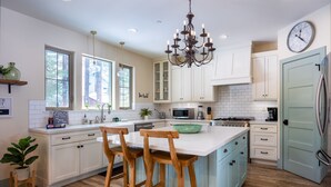 Een moderne keuken met witte kasten, een centraal eiland met barkrukken, een kroonluchter, een klok en een koelkast.