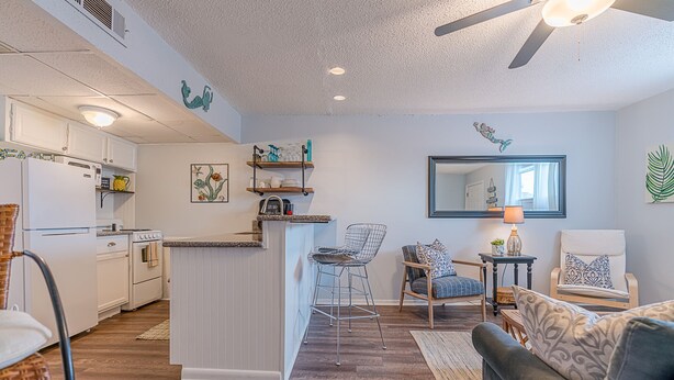 A modern kitchen with white appliances, a dining area with a gray sofa and patterned cushions, and a living room with a large mirror and decorative wall art.