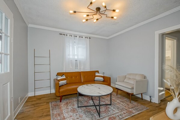 Una sala de estar con un sofá marrón, un sillón blanco, una mesa de centro redonda y un tapete estampado.