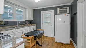 Una cocina con armarios blancos, encimera de granito, una mesa negra, sillas de madera, un refrigerador blanco y una ventana con persianas.