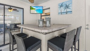 Une terrasse couverte avec une table et des chaises, un ventilateur de plafond, un téléviseur et un tableau représentant des palmiers.