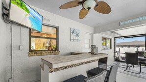Un espace bar aménagé sur le thème de la plage, avec un téléviseur, un ventilateur de plafond et des sièges à l’extérieur.