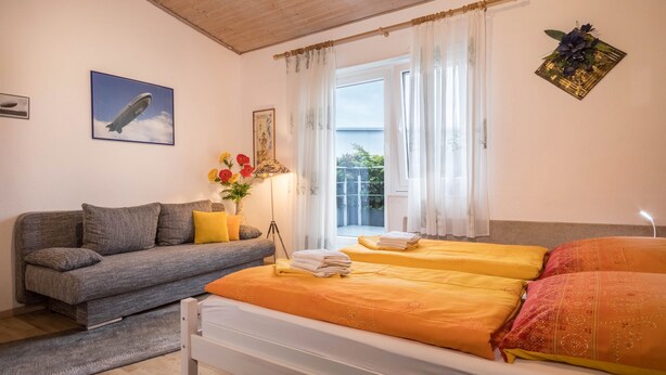 A bedroom with a bed, a sofa, a window with curtains, a picture of a blimp, and a plant. - Studio Apartment 'B + E Röck' with Private Balcony and Wi-Fi (Meckenbeuren)