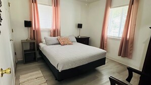 Un dormitorio con una cama, un ventilador de techo, dos mesitas de noche, una silla y una ventana con cortinas.