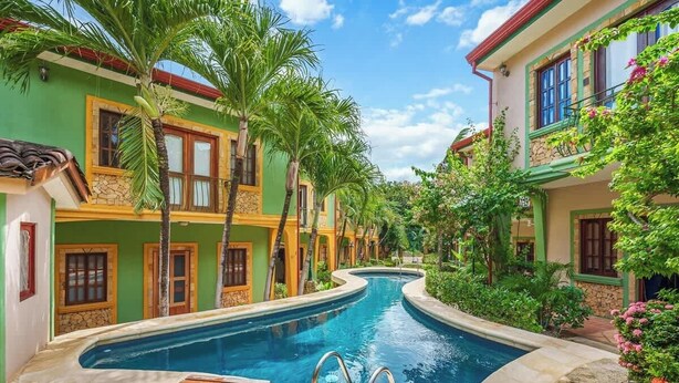 A pool area with a wooden deck, surrounded by buildings with green and yellow facades, and palm trees. - Quiet Tamarindo 2BR | 10-min Walk to the Beach! (Tamarindo)