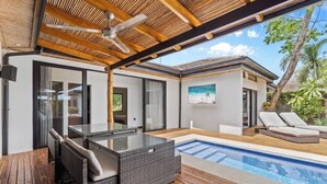 Ein moderner Poolbereich im Freien mit einem Tisch mit Glasplatte und Korbstühlen, Holzdeckböden und einem Deckenventilator.