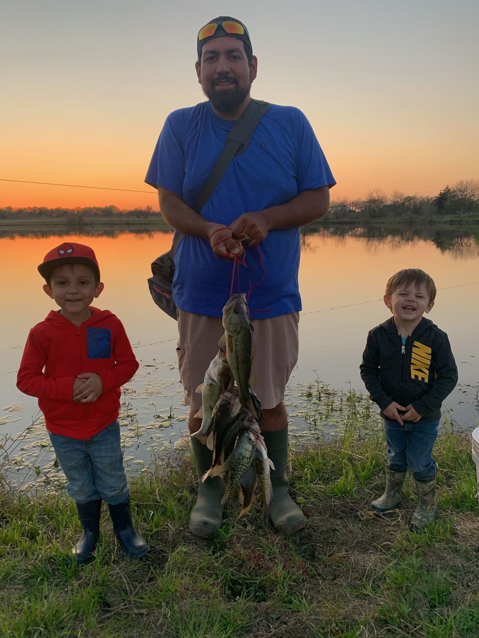 A man holding several fish, standing on grass near a body of water with a sunset in the background.