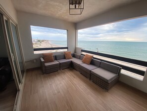 Een kamer met een groot raam dat uitkijkt op een strand, een rieten bank met kussens en een houten vloer.