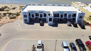 Ein mehrstöckiges Gebäude mit flachem Dach, ein Parkplatz mit mehreren Autos und ein Strand im Hintergrund.