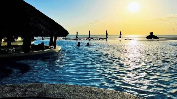 Ein Strand mit einer strohgedeckten Struktur, einem Pool und einem Steg, der ins Wasser führt.