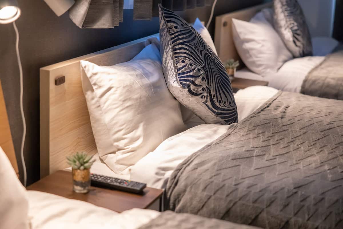 Una habitación de hotel con cabecera de madera, una mesita de noche con un control remoto y una planta, y una cama con ropa de cama estampada.