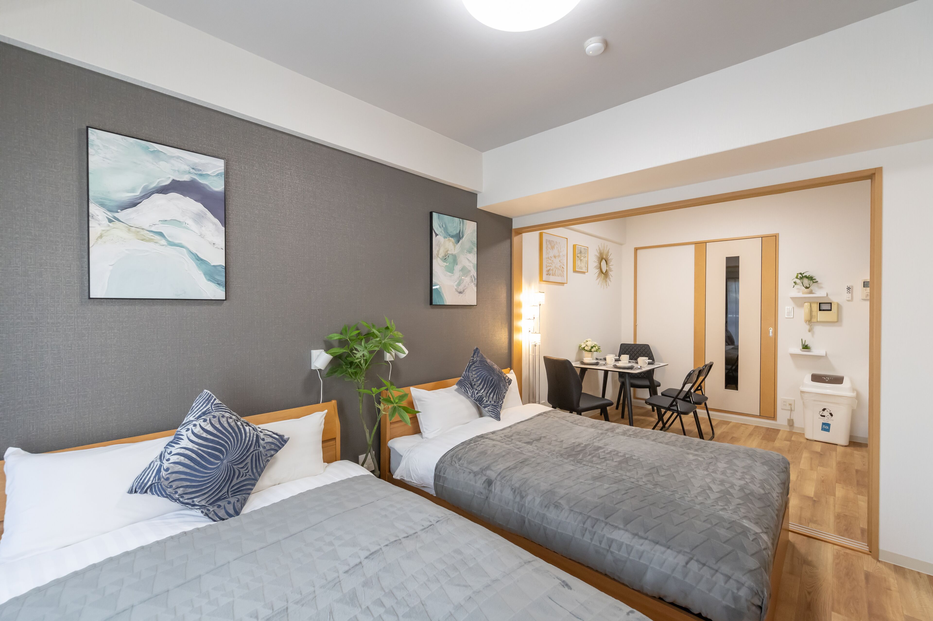 A hotel room with two beds, a desk, and a chair. There are two paintings on the wall, a plant, and a small table with a lamp.