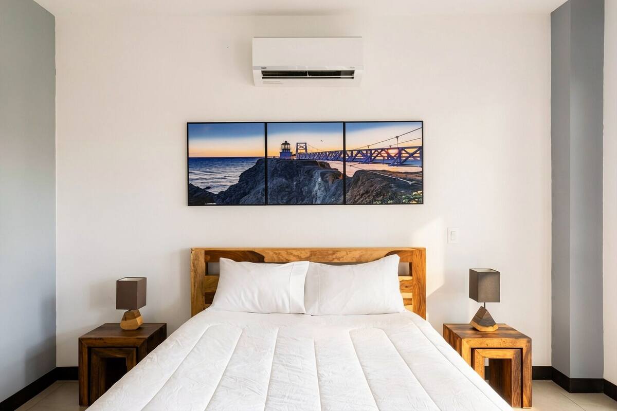 Un dormitorio con cabecera de madera, una cama grande con ropa blanca, dos mesitas de noche con lámparas y un aire acondicionado instalado en la pared.
