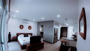 A modern hotel room with a bed, a TV, a sofa, a kitchen area, and a bathroom.