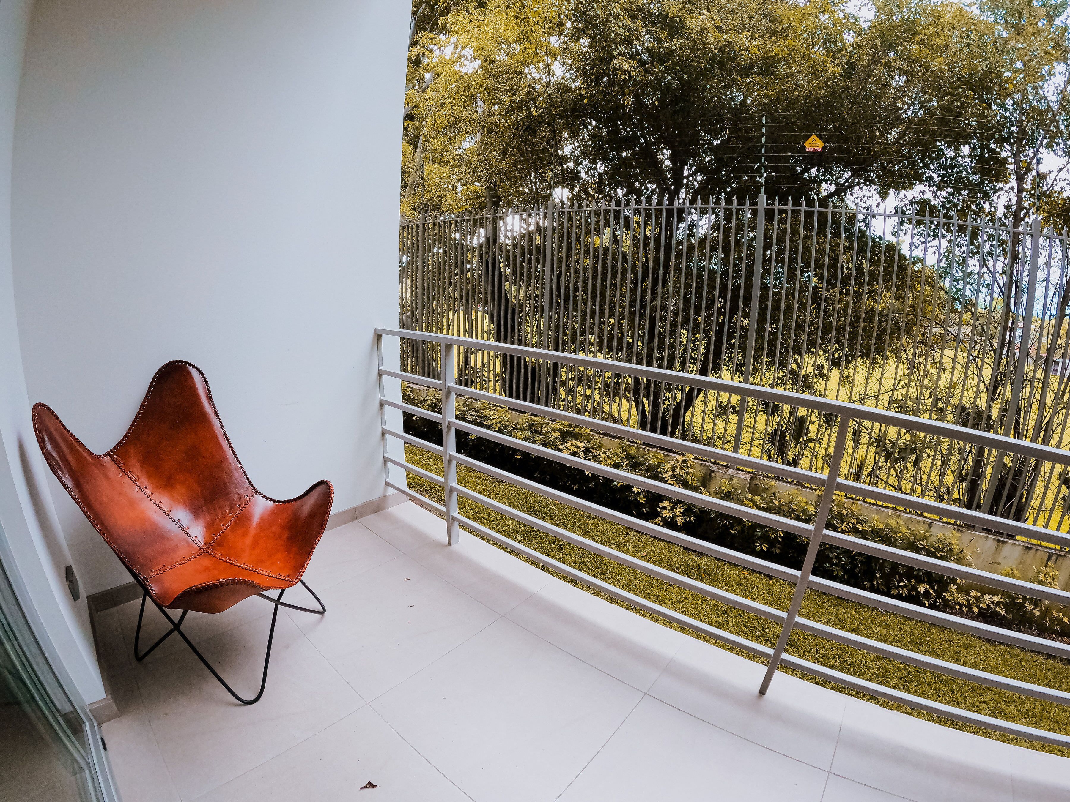 Un balcón con barandilla blanca, un sillón de cuero marrón y vistas a árboles y una cerca.