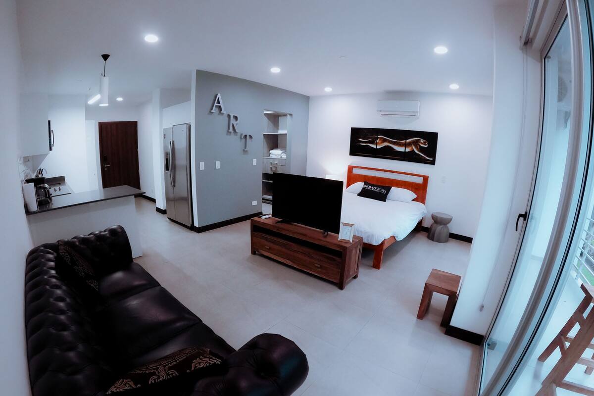 Una sala de estar moderna con un sofá de cuero negro, una mesa de centro de madera y un televisor sobre un mueble bajo. Se observa una cama con cabecera blanca y un cuadro sobre ella. En el fondo, se distingue una zona de cocina con un refrigerador y un lavamanos.