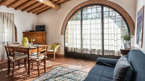 Un salon chaleureux avec une table à manger en bois, des chaises, un canapé et une grande fenêtre en arc surmontée de rideaux.