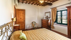 Une chambre à coucher avec un lit en bois, une commode en bois, une chaise, une fenêtre avec des rideaux et un plafond avec des poutres apparentes.