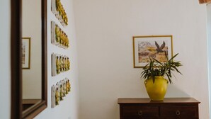 Une pièce avec un miroir qui reflète une œuvre d’art accrochée au mur, un tableau encadré et un vase jaune posé sur une commode en bois.