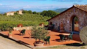 Ein Steinhaus mit einer gefliesten Terrasse, Sitzgelegenheiten im Freien und Blick auf Weinberge und Hügel.