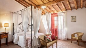 Ein Schlafzimmer mit Himmelbett, Korbsessel, Nachttisch und rotem Vorhang.