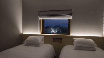 A hotel room with two beds, a window with a view of a cityscape at night, and a wall-mounted control panel.