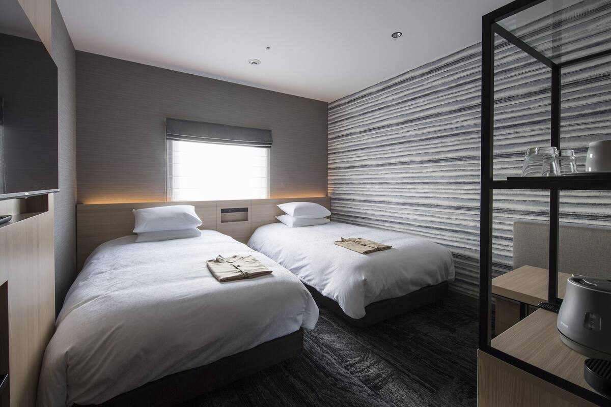 A modern hotel room with two beds, a large window, a flat-screen TV, a minibar, and a large mirror.
