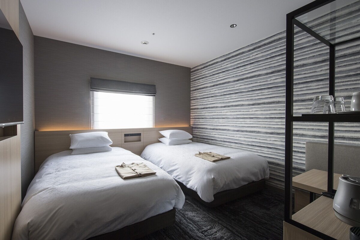 A hotel room with two beds, a large window, a modern wall design, and a glass partition with a kettle.