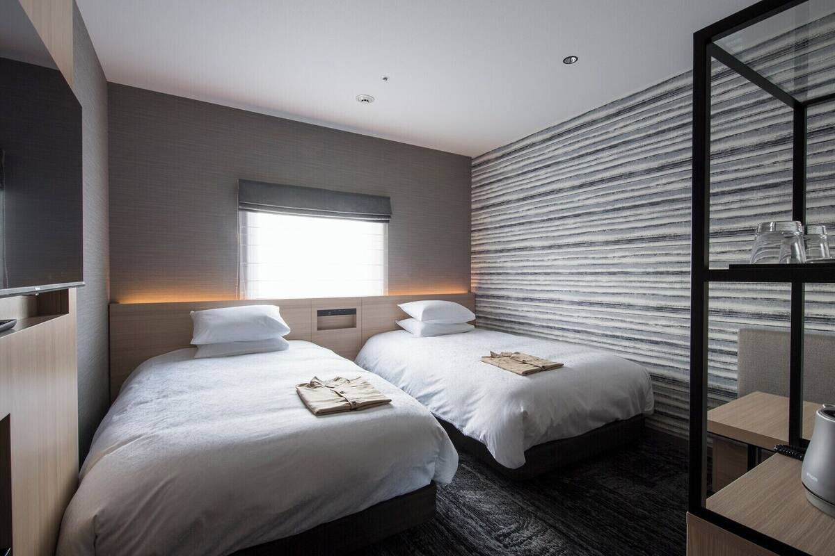 A hotel room with two beds, a large window, a wall with a textured pattern, and a glass partition with a kettle and cups.