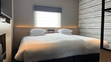 A hotel room with a bed, a window with blinds, a bedside table, and a wall-mounted TV.