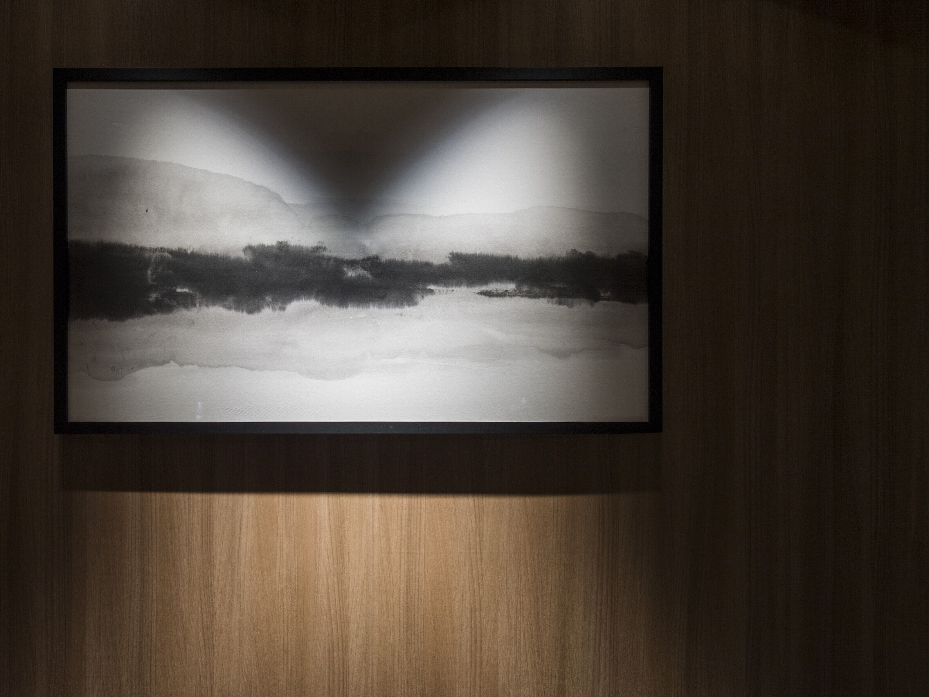 Fotografía en blanco y negro enmarcada de un paisaje con una cadena montañosa y una masa de agua, expuesta en una pared de madera.