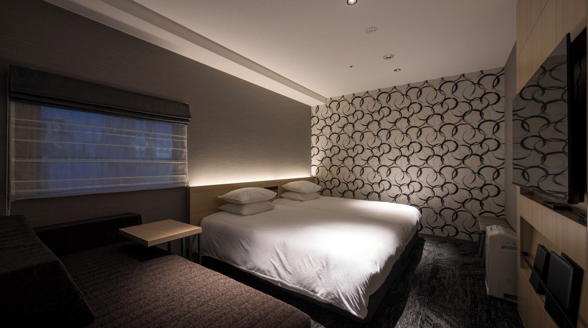 Een moderne hotelkamer met een groot bed, een nachtkastje, een flatscreen televisie aan de muur en een raam met zonwering.