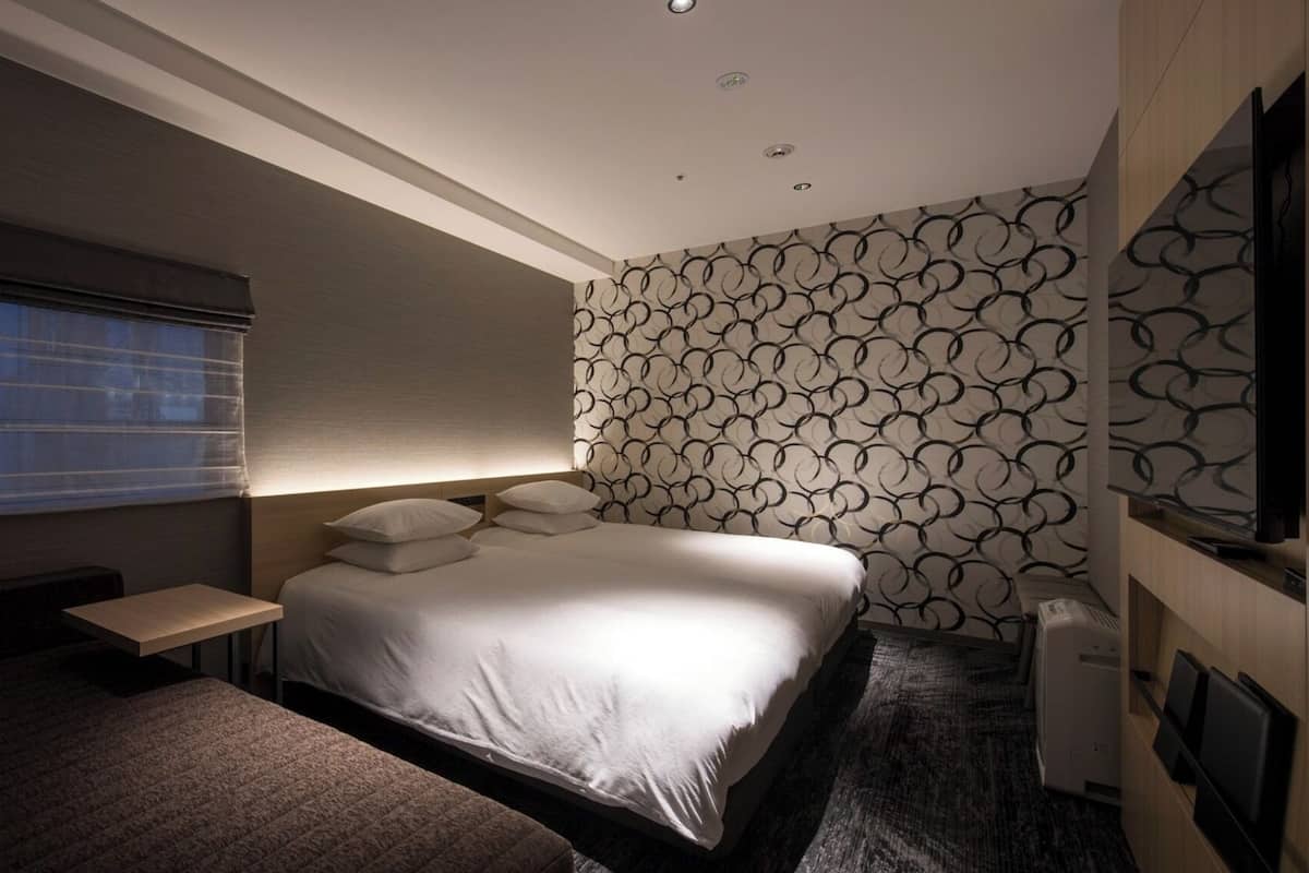 Une chambre d’hôtel moderne dotée d’un grand lit, d’une table de chevet, d’un téléviseur à écran plat fixé au mur et d’une fenêtre avec des stores.