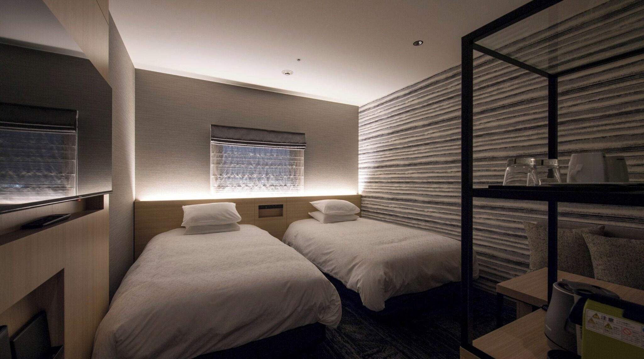 Hotelkamer met twee eenpersoonsbedden, een raam met zonwering, een inbouw bureau en een wand met een structuurpatroon.