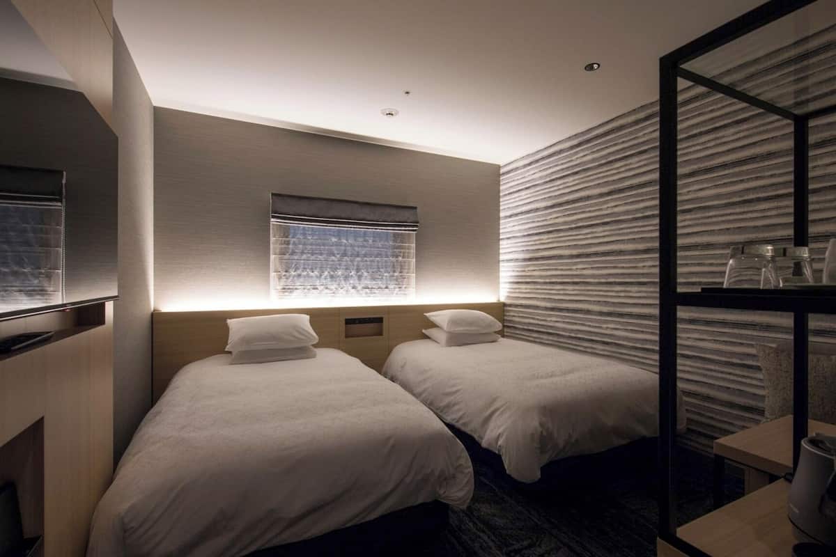 Une chambre d’hôtel avec deux lits simples, une fenêtre avec des stores, un bureau intégré et un mur à motifs.