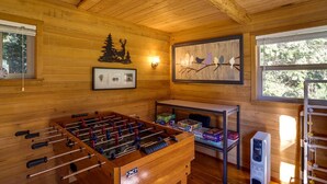 Une pièce au plancher de bois, avec un baby-foot, un tableau représentant des oiseaux perchés sur une branche et une fenêtre donnant sur des arbres.