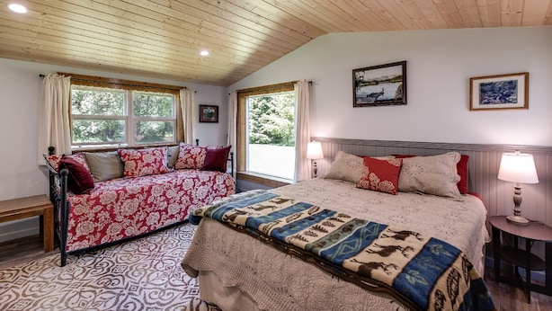 Une chambre avec un plafond en bois, un grand lit avec une literie à motifs, un canapé recouvert d’un tissu à fleurs et deux fenêtres avec des rideaux.
