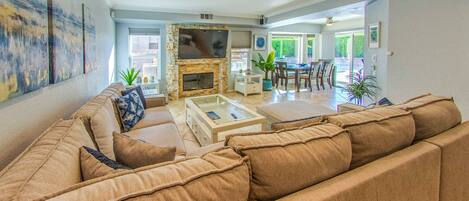 A living room with a large beige sectional sofa, a glass coffee table, a flat-screen TV mounted on a stone fireplace, and a dining area with a table and chairs.